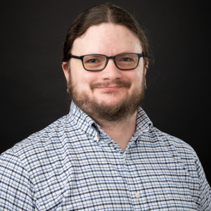 Studio portrait of a bearded man with long brown hair and black glasses, wearing a blue-and-white checked shirt against a dark background, smiling gently.