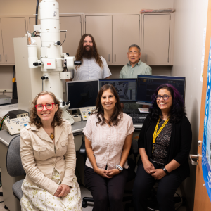 Five researchers pose in a laboratory beside a large microscope and computer monitors, smiling warmly in a collaborative, welcoming setting.